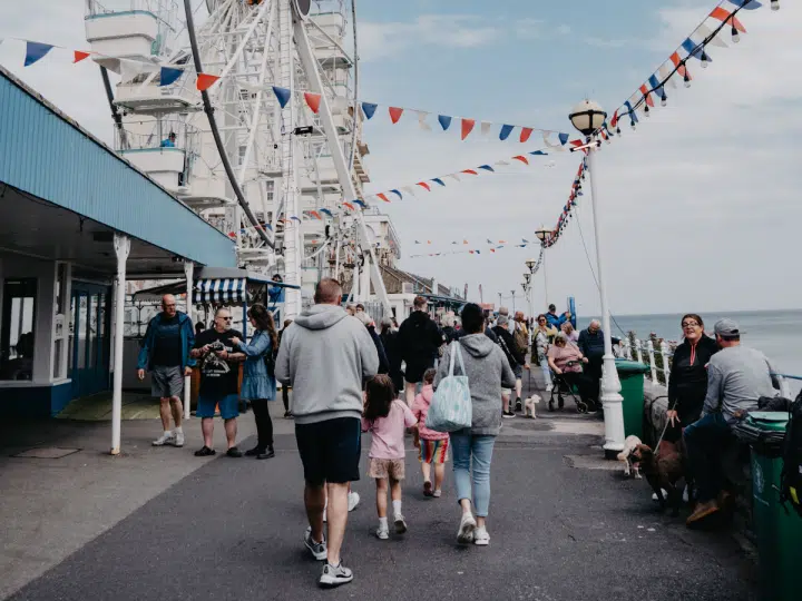 A bustling scene at Llandudno promenade in Wales with people walking near a Ferris wheel adorned with flags. Days out like this will not be affordable for many UK families due to money worries.