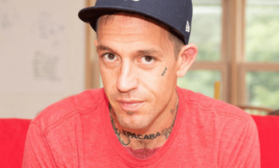Fergie Chambers, who has small face tattoos and a neck tattoo on show above his crew neck tshirt, looks at the camera from beneath his baseball cap. He is a vocal supporter of Palestine.
