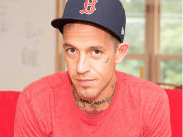 Fergie Chambers, who has small face tattoos and a neck tattoo on show above his crew neck tshirt, looks at the camera from beneath his baseball cap. He is a vocal supporter of Palestine.