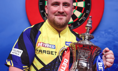 Luke "the Nuke" Littler smiles with his winner's trophy in hand infront of a darts board in Blackpool