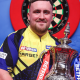 Luke "the Nuke" Littler smiles with his winner's trophy in hand infront of a darts board in Blackpool