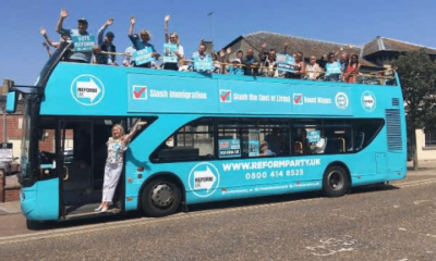 A Reform branded open top double decker bus parked with people on top waving and a woman in the doorway waving too, in 2013