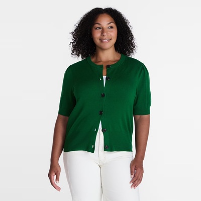 A woman wearing a green short-sleeved cardigan, white top, and white pants