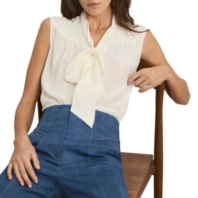 A woman sitting on a chair, wearing an ivory tie-neck blouse and high-waisted blue jeans