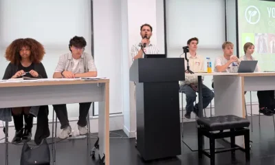 Young Greens party conference. Each candidate is sat at the front of a classroom on two desks with a podium and mic in the middle, where one candidate is speaking
