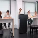 Young Greens party conference. Each candidate is sat at the front of a classroom on two desks with a podium and mic in the middle, where one candidate is speaking