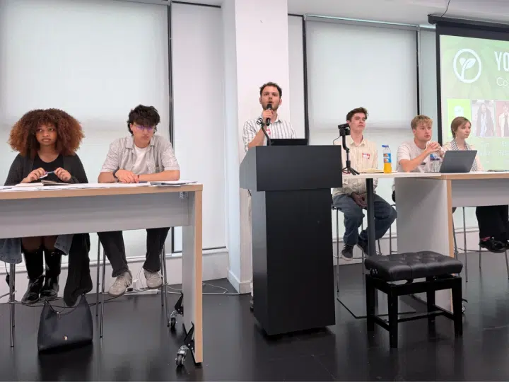 Young Greens party conference. Each candidate is sat at the front of a classroom on two desks with a podium and mic in the middle, where one candidate is speaking