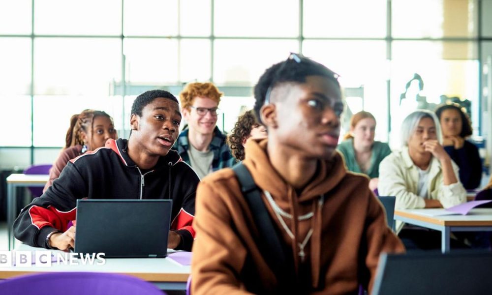 Students sit at desks with laptops in a bright classroom or lecture space. Several people are facing forward as if listening to a lesson, with large windows letting in daylight behind them.