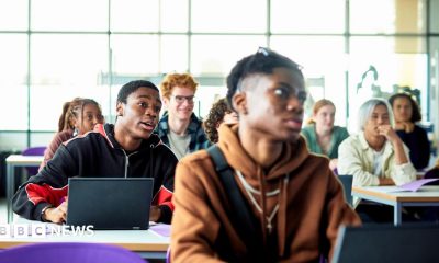 Students sit at desks with laptops in a bright classroom or lecture space. Several people are facing forward as if listening to a lesson, with large windows letting in daylight behind them.