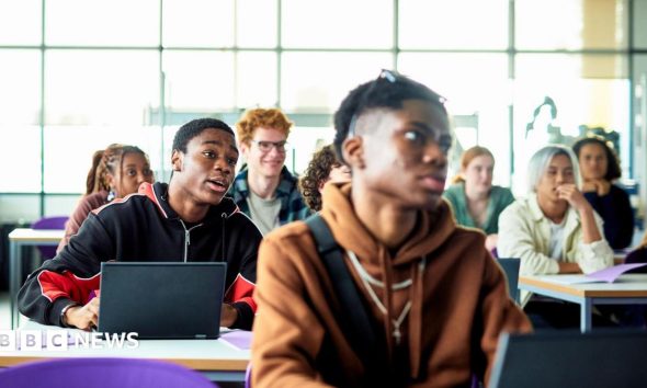 Students sit at desks with laptops in a bright classroom or lecture space. Several people are facing forward as if listening to a lesson, with large windows letting in daylight behind them.