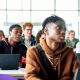 Students sit at desks with laptops in a bright classroom or lecture space. Several people are facing forward as if listening to a lesson, with large windows letting in daylight behind them.