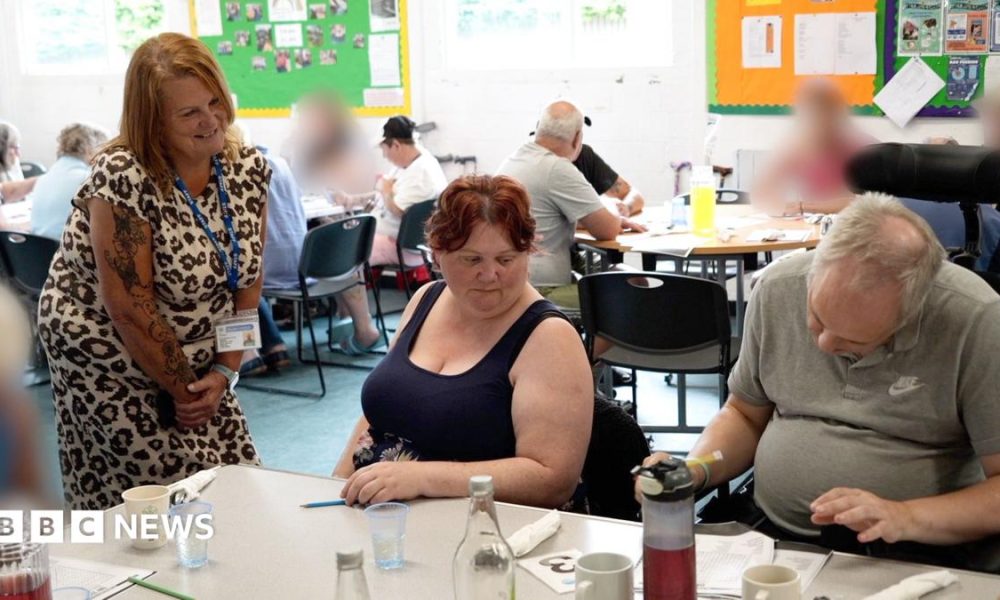 A woman wearing a leopard-print dress bends at a table where a man and woman sit. The man is filling out a form. There are mugs on the table with bottles of water. In the background more people sit around tables.