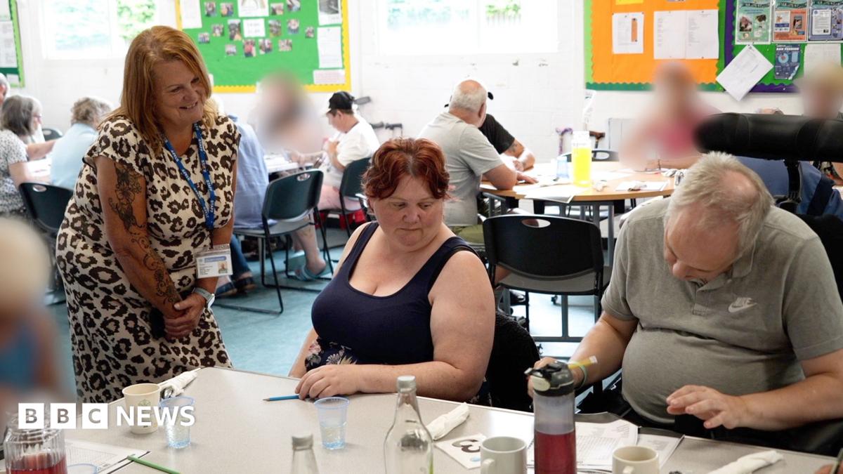 A woman wearing a leopard-print dress bends at a table where a man and woman sit. The man is filling out a form. There are mugs on the table with bottles of water. In the background more people sit around tables.