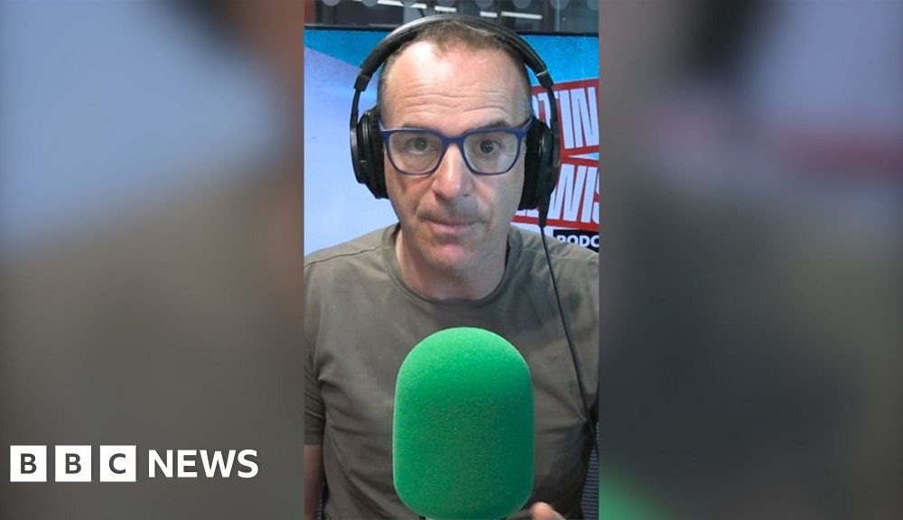 Martin Lewis is wearing dark green T-shirt and black-framed glasses. He is wearing headphones and sitting behind a green mic.