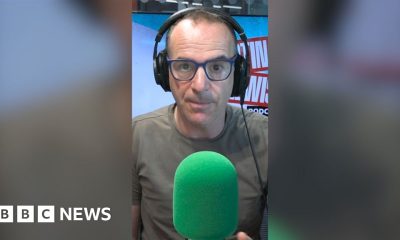 Martin Lewis is wearing dark green T-shirt and black-framed glasses. He is wearing headphones and sitting behind a green mic.
