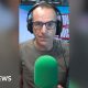 Martin Lewis is wearing dark green T-shirt and black-framed glasses. He is wearing headphones and sitting behind a green mic.