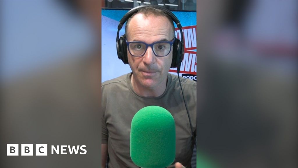 Martin Lewis is wearing dark green T-shirt and black-framed glasses. He is wearing headphones and sitting behind a green mic.