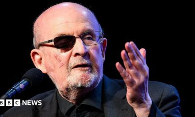 Rushdie speaking in March 2026 into a microphone. He has a darkened lens over his wounded eye