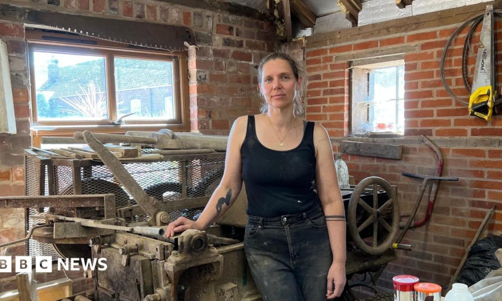 Tessa stands inside a workshop, resting one hand on a large, old industrial machine with metal rollers and levers. She is wearing a black sleeveless vest and black, dust-marked jeans. The workshop has exposed red brick walls, wooden beams, and two small rectangular windows behind her which are letting in daylight. Around the machine are various tools and materials, including wooden planks, metal parts, and a saw mounted on the wall to the left.
