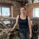 Tessa stands inside a workshop, resting one hand on a large, old industrial machine with metal rollers and levers. She is wearing a black sleeveless vest and black, dust-marked jeans. The workshop has exposed red brick walls, wooden beams, and two small rectangular windows behind her which are letting in daylight. Around the machine are various tools and materials, including wooden planks, metal parts, and a saw mounted on the wall to the left.