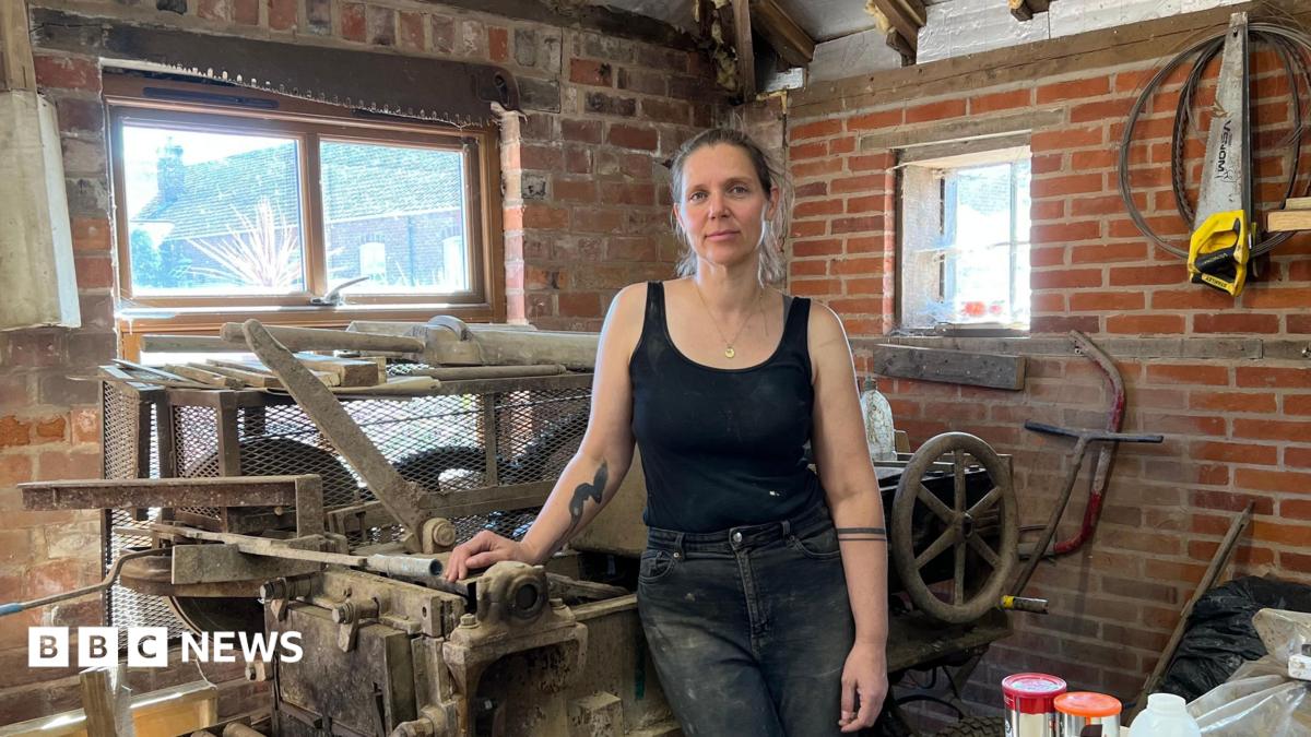 Tessa stands inside a workshop, resting one hand on a large, old industrial machine with metal rollers and levers. She is wearing a black sleeveless vest and black, dust-marked jeans. The workshop has exposed red brick walls, wooden beams, and two small rectangular windows behind her which are letting in daylight. Around the machine are various tools and materials, including wooden planks, metal parts, and a saw mounted on the wall to the left.