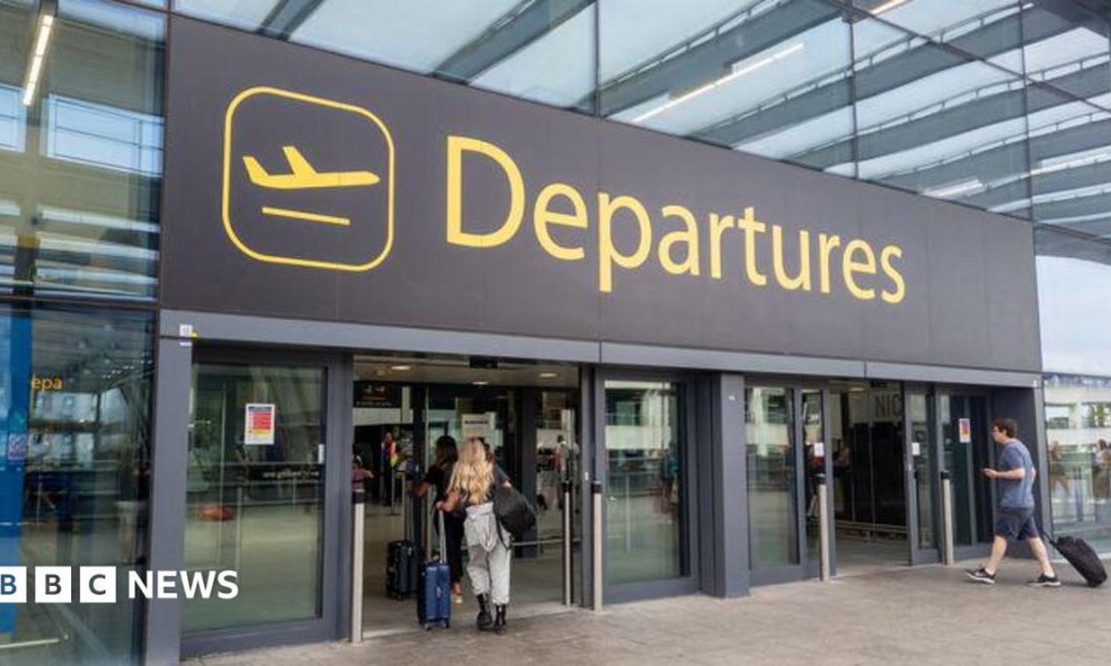 The departures entrance at Gatwick Airport with large yellow "Departures" signage above glass doors. Travellers with luggage can be seen entering the terminal building.