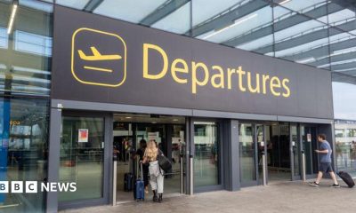 The departures entrance at Gatwick Airport with large yellow "Departures" signage above glass doors. Travellers with luggage can be seen entering the terminal building.