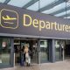 The departures entrance at Gatwick Airport with large yellow "Departures" signage above glass doors. Travellers with luggage can be seen entering the terminal building.