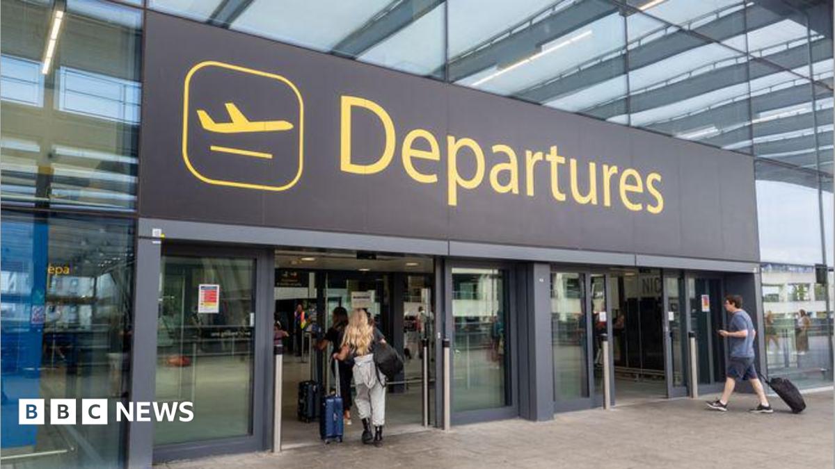 The departures entrance at Gatwick Airport with large yellow "Departures" signage above glass doors. Travellers with luggage can be seen entering the terminal building.
