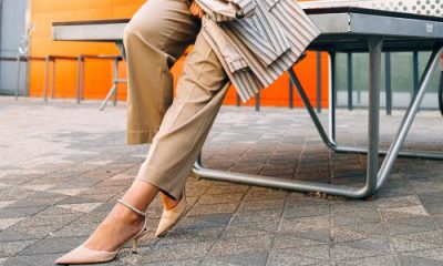 professional young woman wears stylish summer work outfit in beige pants, nude heels, white blouse and striped blazer; she is leaning against a table outside on cobblestones; in the background is an orange wall