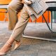 professional young woman wears stylish summer work outfit in beige pants, nude heels, white blouse and striped blazer; she is leaning against a table outside on cobblestones; in the background is an orange wall