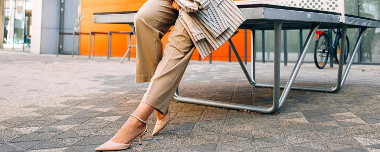 professional young woman wears stylish summer work outfit in beige pants, nude heels, white blouse and striped blazer; she is leaning against a table outside on cobblestones; in the background is an orange wall