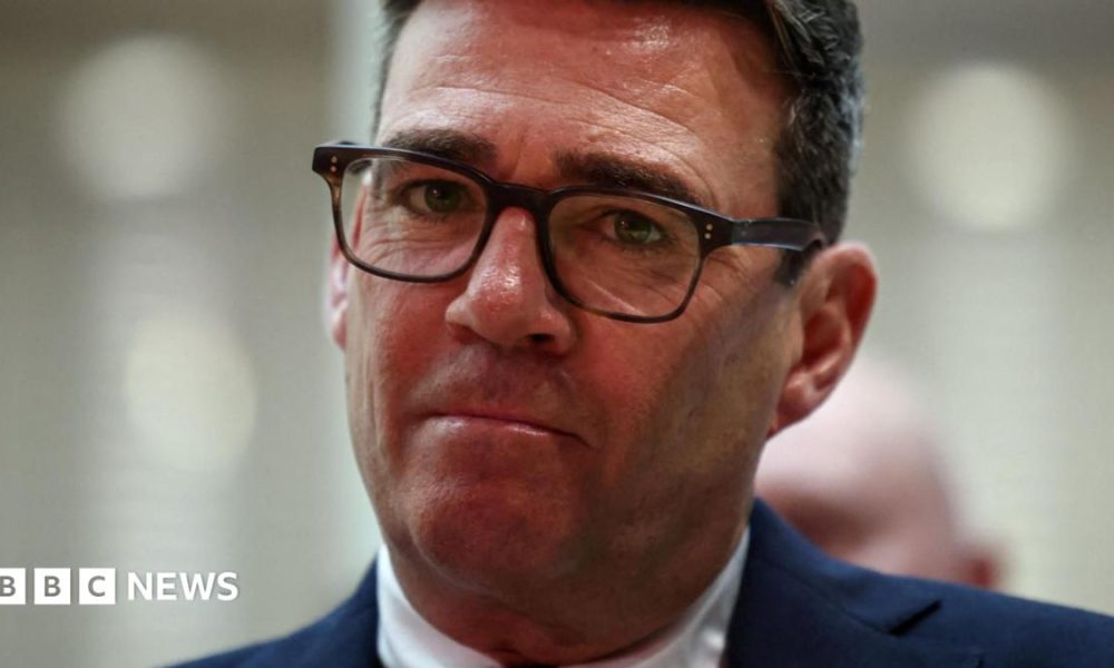 Andy Burnham looks directly forward, his lips are purse and he wears tortoise shell glasses. His suit jacket is navy and his shirt is white. The background is blurred grey.