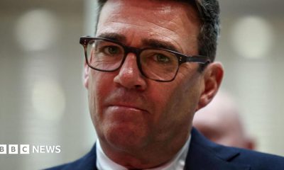 Andy Burnham looks directly forward, his lips are purse and he wears tortoise shell glasses. His suit jacket is navy and his shirt is white. The background is blurred grey.