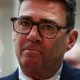Andy Burnham looks directly forward, his lips are purse and he wears tortoise shell glasses. His suit jacket is navy and his shirt is white. The background is blurred grey.