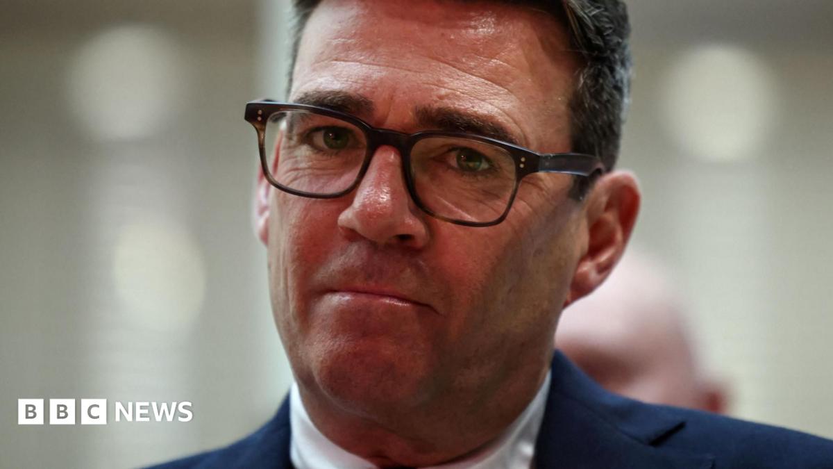 Andy Burnham looks directly forward, his lips are purse and he wears tortoise shell glasses. His suit jacket is navy and his shirt is white. The background is blurred grey.