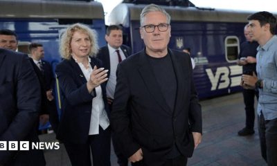 Sir Keir Starmer is walking on a train platform. A blue-coloured train is in the background. Sir Keir is wearing a dark-coloured jacket, top and trousers. There are other people on the platform to his left and behind him.
