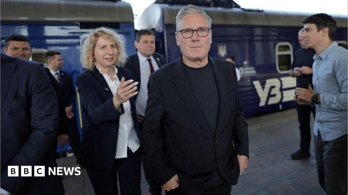 Sir Keir Starmer is walking on a train platform. A blue-coloured train is in the background. Sir Keir is wearing a dark-coloured jacket, top and trousers. There are other people on the platform to his left and behind him.