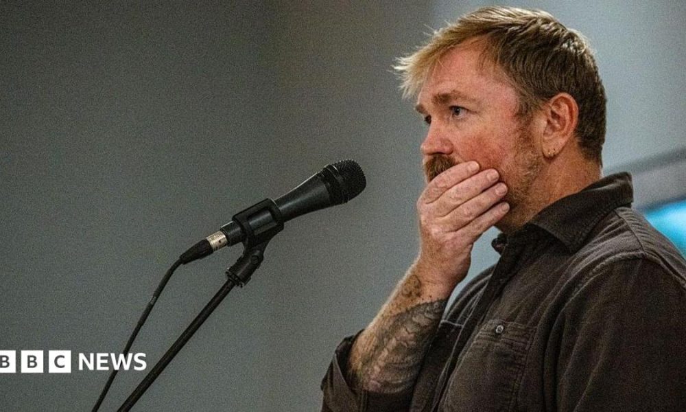 Graham Platner seen on stage at a microphone. his hand is over his mouth