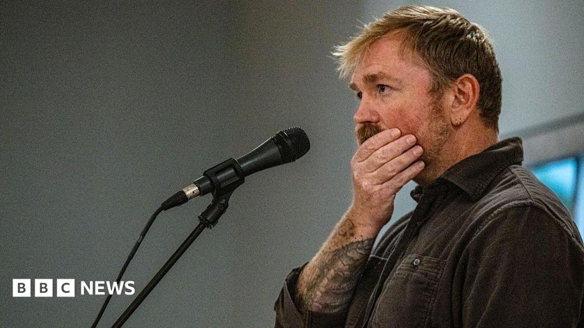 Graham Platner seen on stage at a microphone. his hand is over his mouth