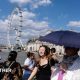 People walk on a bridge over the Thames in London using an umbrella for shade with strong sunshine overhead.