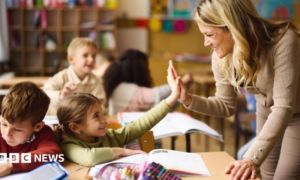 A young teacher is leaning over the desk of two under-10 pupils and is high-fiving the nearest child. Both are smiling.