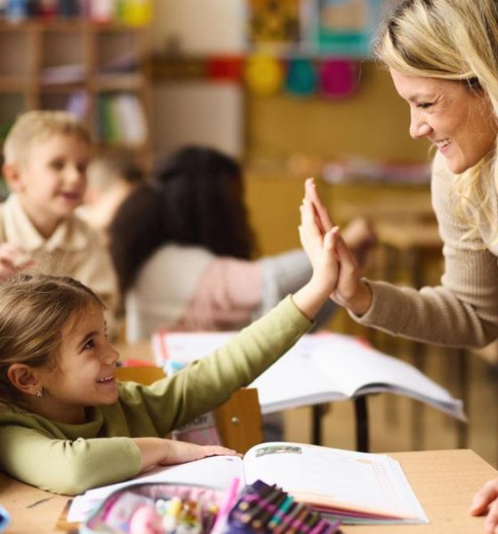 A young teacher is leaning over the desk of two under-10 pupils and is high-fiving the nearest child. Both are smiling.