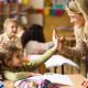 A young teacher is leaning over the desk of two under-10 pupils and is high-fiving the nearest child. Both are smiling.