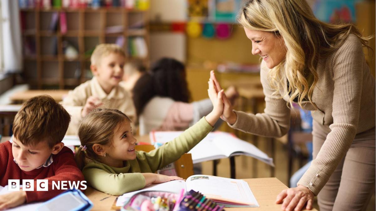 A young teacher is leaning over the desk of two under-10 pupils and is high-fiving the nearest child. Both are smiling.