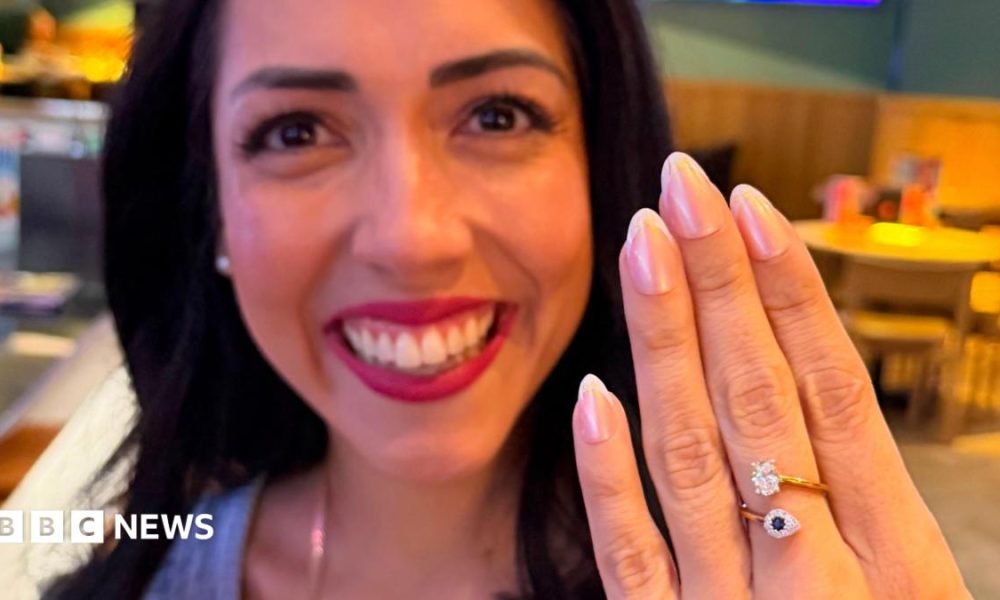 A close up of a young woman smiling broadly and holding her hand up in front of  her. She has a ring on her middle finger with a sapphire and diamond.