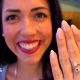 A close up of a young woman smiling broadly and holding her hand up in front of  her. She has a ring on her middle finger with a sapphire and diamond.