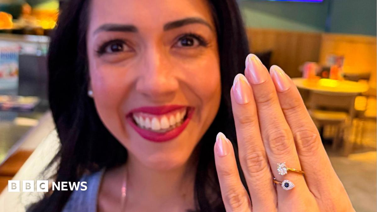 A close up of a young woman smiling broadly and holding her hand up in front of  her. She has a ring on her middle finger with a sapphire and diamond.