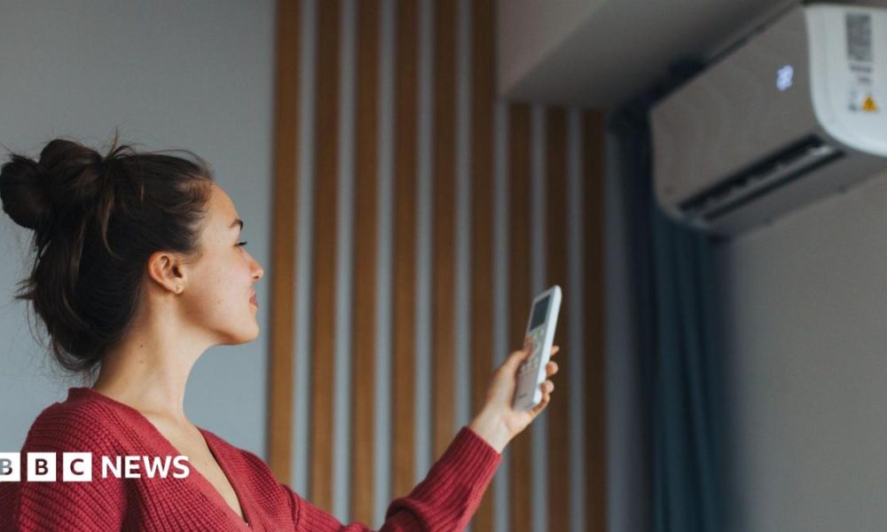 A woman with brown hair, wearing a red cardigan, is turning on a wall-mounted air-conditioning unit with a remote.