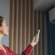 A woman with brown hair, wearing a red cardigan, is turning on a wall-mounted air-conditioning unit with a remote.